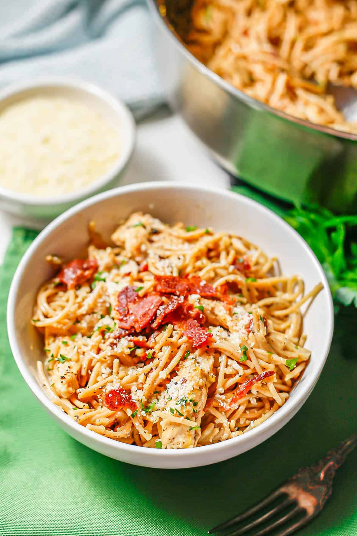 A serving of chicken carbonara in a round white bowl with bacon, parsley and Parmesan on top and the skillet in the background.