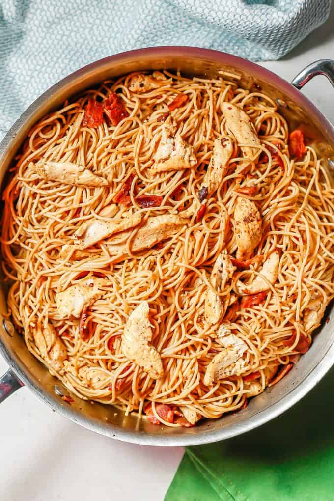 A deep All Clad skillet filled with spaghetti noodles, chicken strips and cooked pieces of bacon.