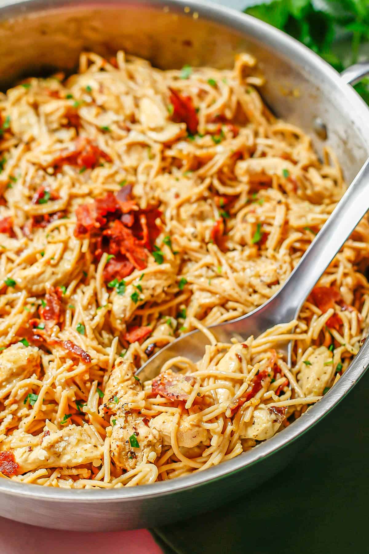 A large silver serving spoon taking a scoop of chicken carbonara from a deep skillet.