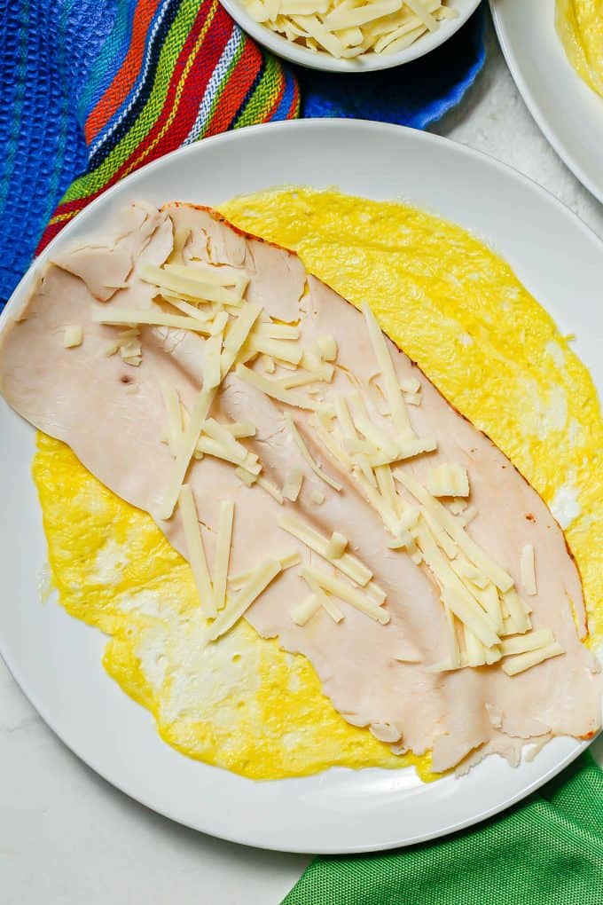 An egg wrap topped with deli turkey and shredded cheese on a white plate.