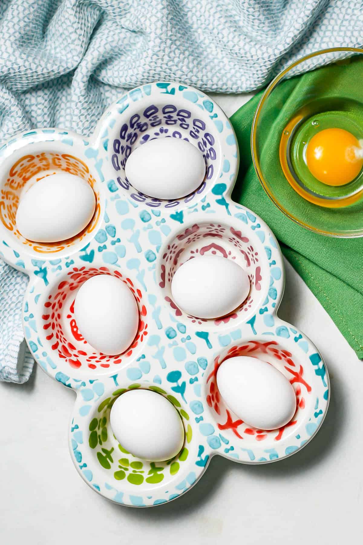 A half dozen eggs in a colorful muffin container with a cracked egg in a bowl to the side.