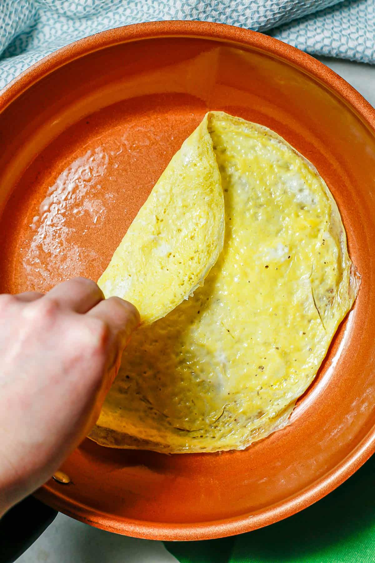 A hand lifting up a thin layer of cooked egg in a copper skillet.