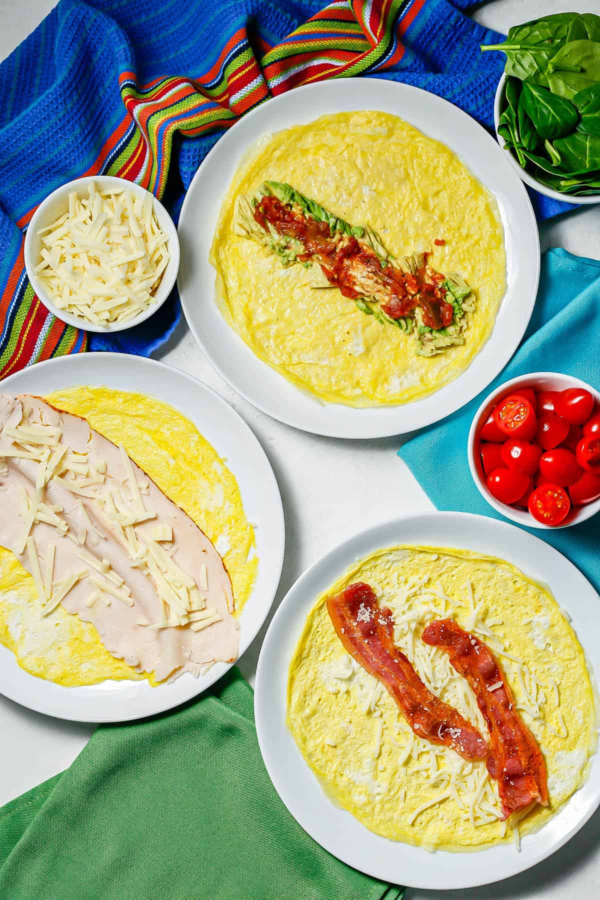 A trio of egg wraps on round white plates with different toppings and extra ingredients in bowls to the side.