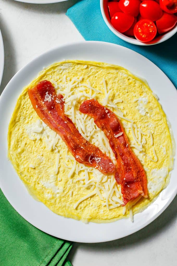 An egg wrap topped with shredded cheese and two cooked slices of bacon.