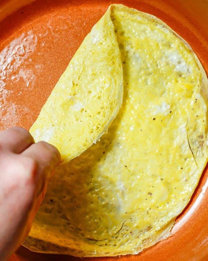 A hand folding over a cooked egg wrap in a copper skillet.