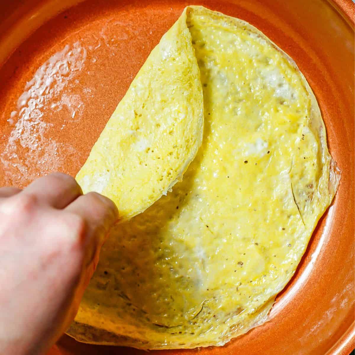 A hand folding over a cooked egg wrap in a copper skillet.