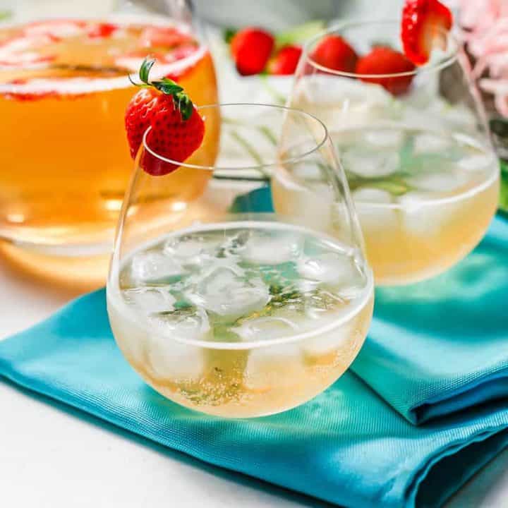 Two stemless wine glasses with an bubbly cocktail garnished with strawberries and a pitcher of the cocktail in the background.