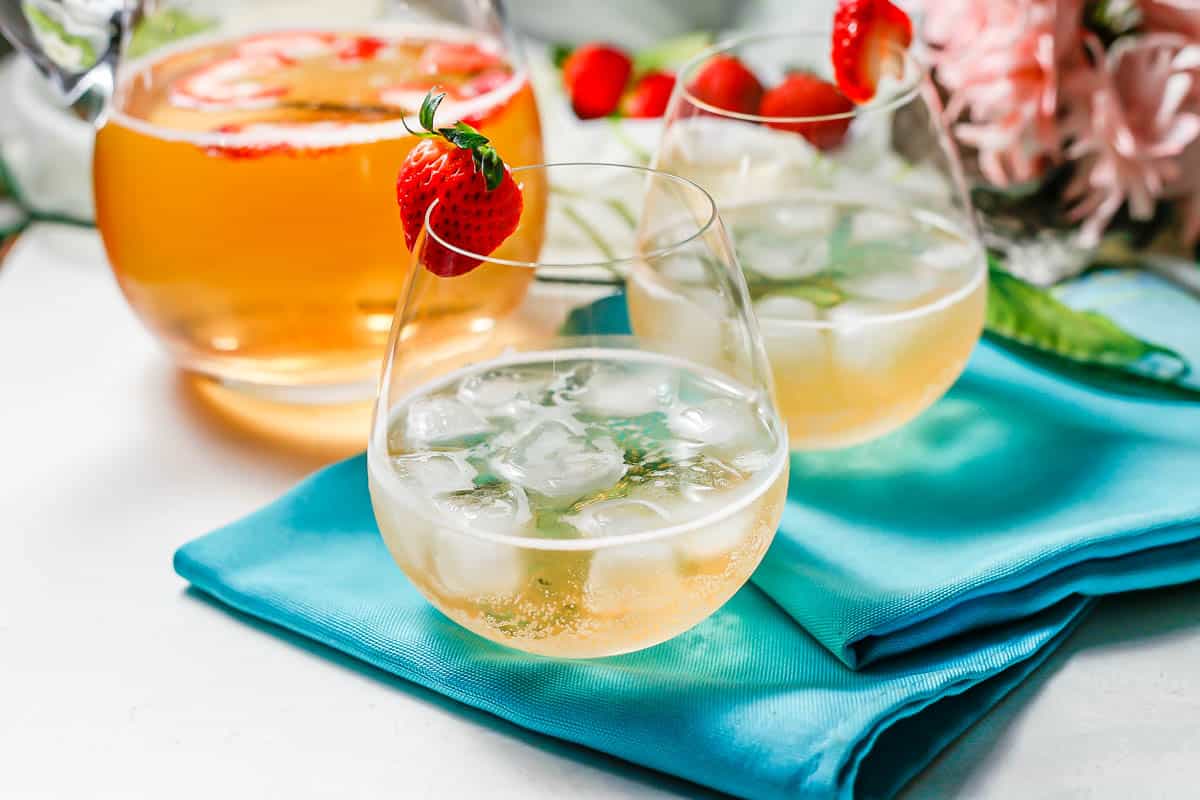 Two stemless wine glasses with an bubbly cocktail garnished with strawberries and a pitcher of the cocktail in the background.