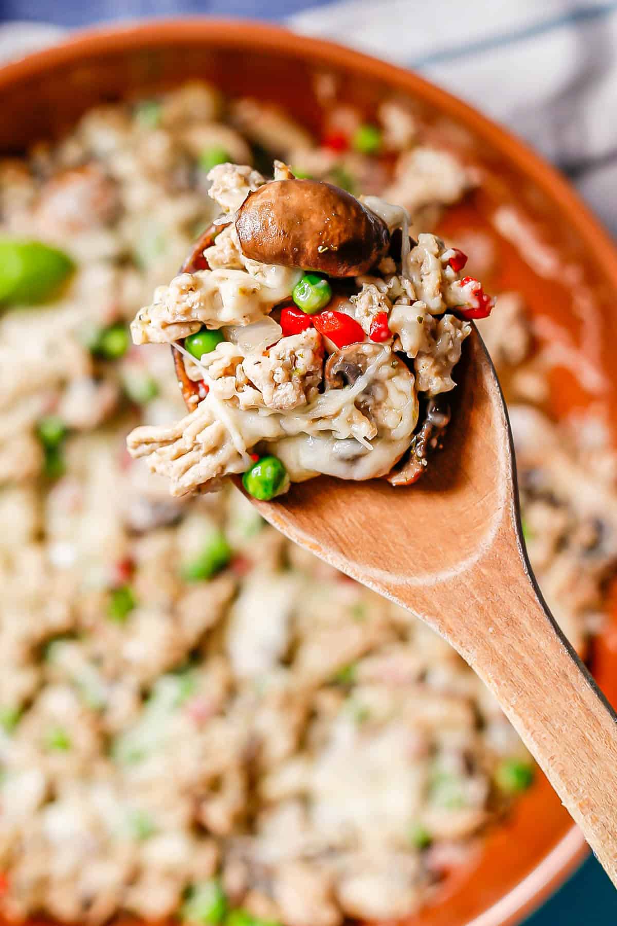 A wooden spoon lifting up a scoop of a ground chicken mixture with mushrooms, peas, pimientos and cheese.