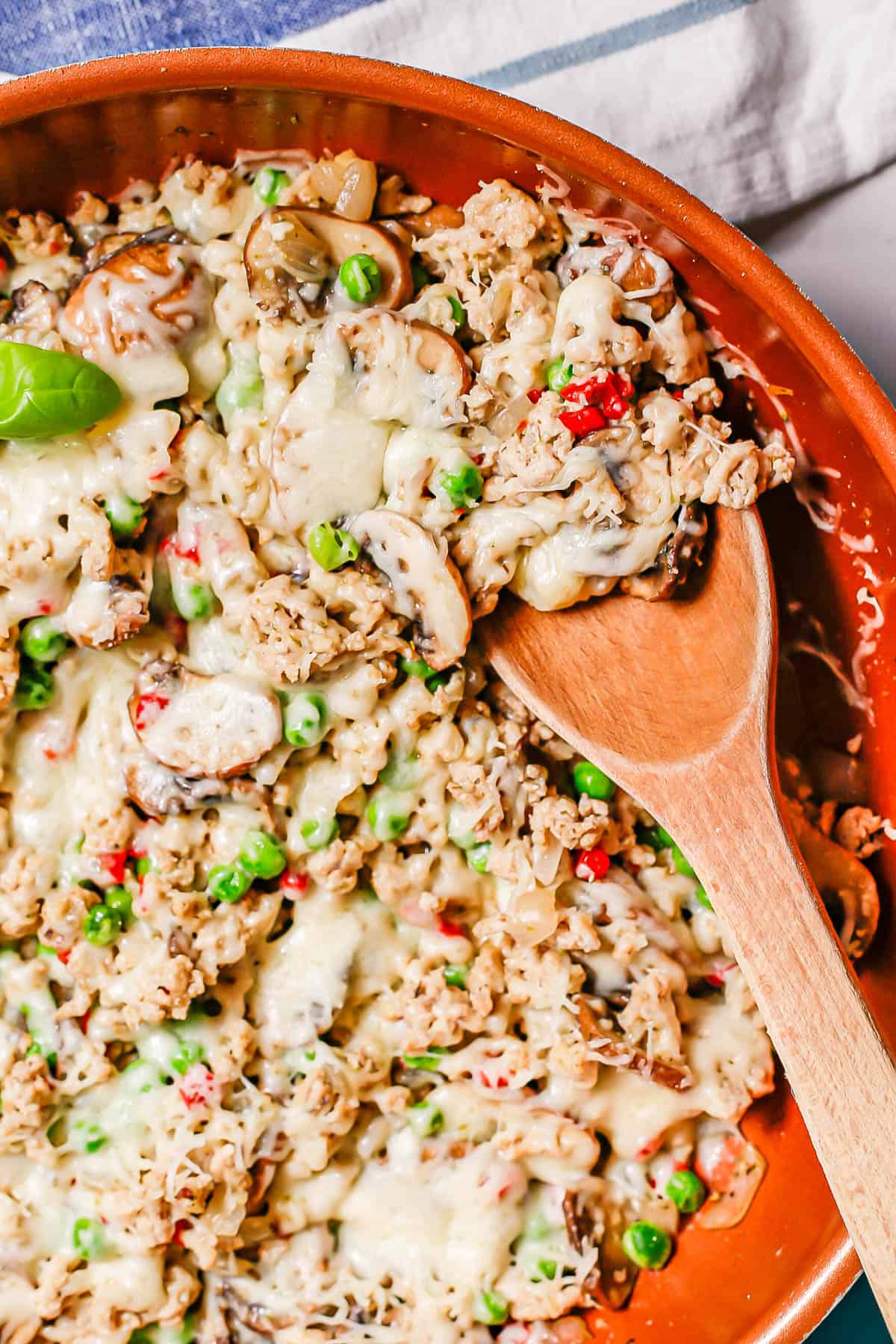 A wooden spoon resting in a copper skillet with a mixture of ground chicken, mushrooms and peas topped with cheese.