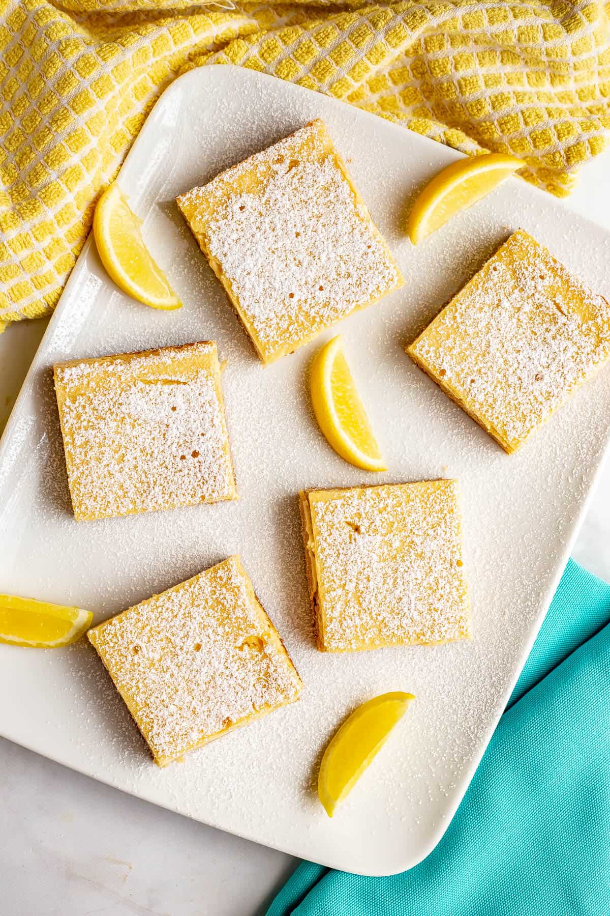 Five lemon bars arranged on a white serving plate with lemon wedges scattered and powdered sugar sprinkled on top.