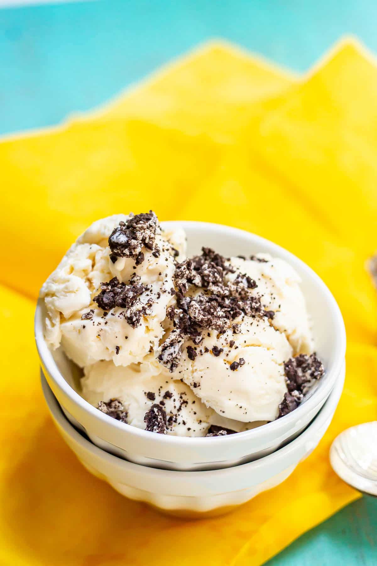 Vanilla ice cream topped with crushed Oreo cookie pieces served in a stacked white bowl on a yellow fabric with spoons nearby.