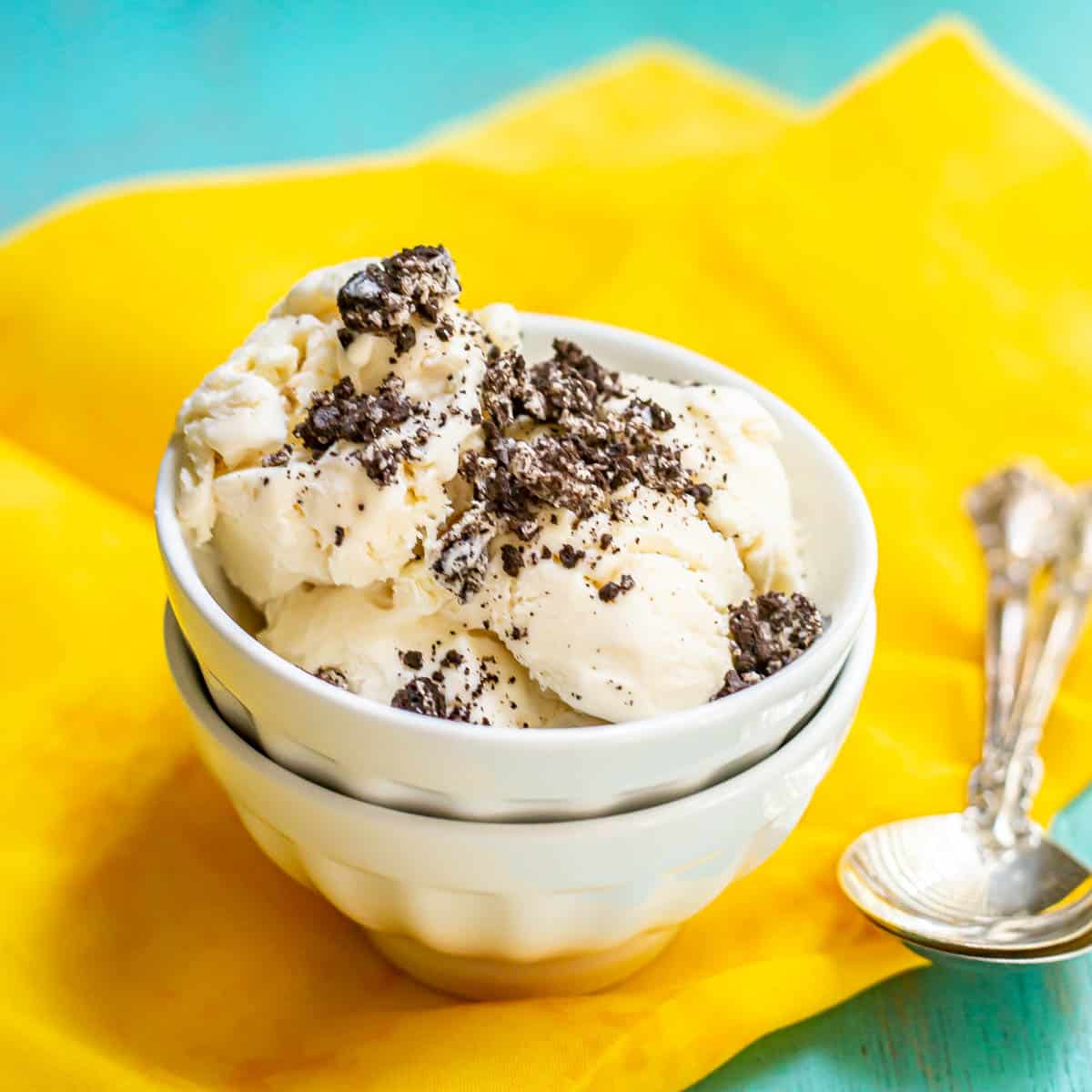 Close up of homemade vanilla no churn ice cream topped with crushed Oreo cookie pieces served in a stacked white bowl on a yellow fabric with spoons nearby.