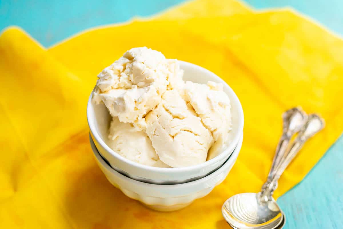 Scoops of vanilla ice cream served in a stacked white bowl on a yellow fabric with spoons nearby.