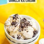 Close up of homemade vanilla no churn ice cream topped with crushed Oreo cookie pieces served in a stacked white bowl on a yellow fabric with spoons nearby and a text overlay on the photo.