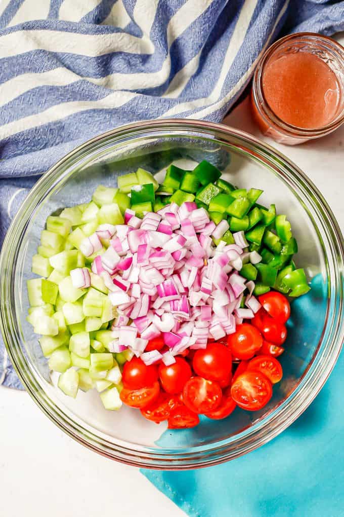 A glass bowl with diced cucumber, green pepper, red onion and tomatoes arranged in separate piles and a red wine dressing in a jar to the side.