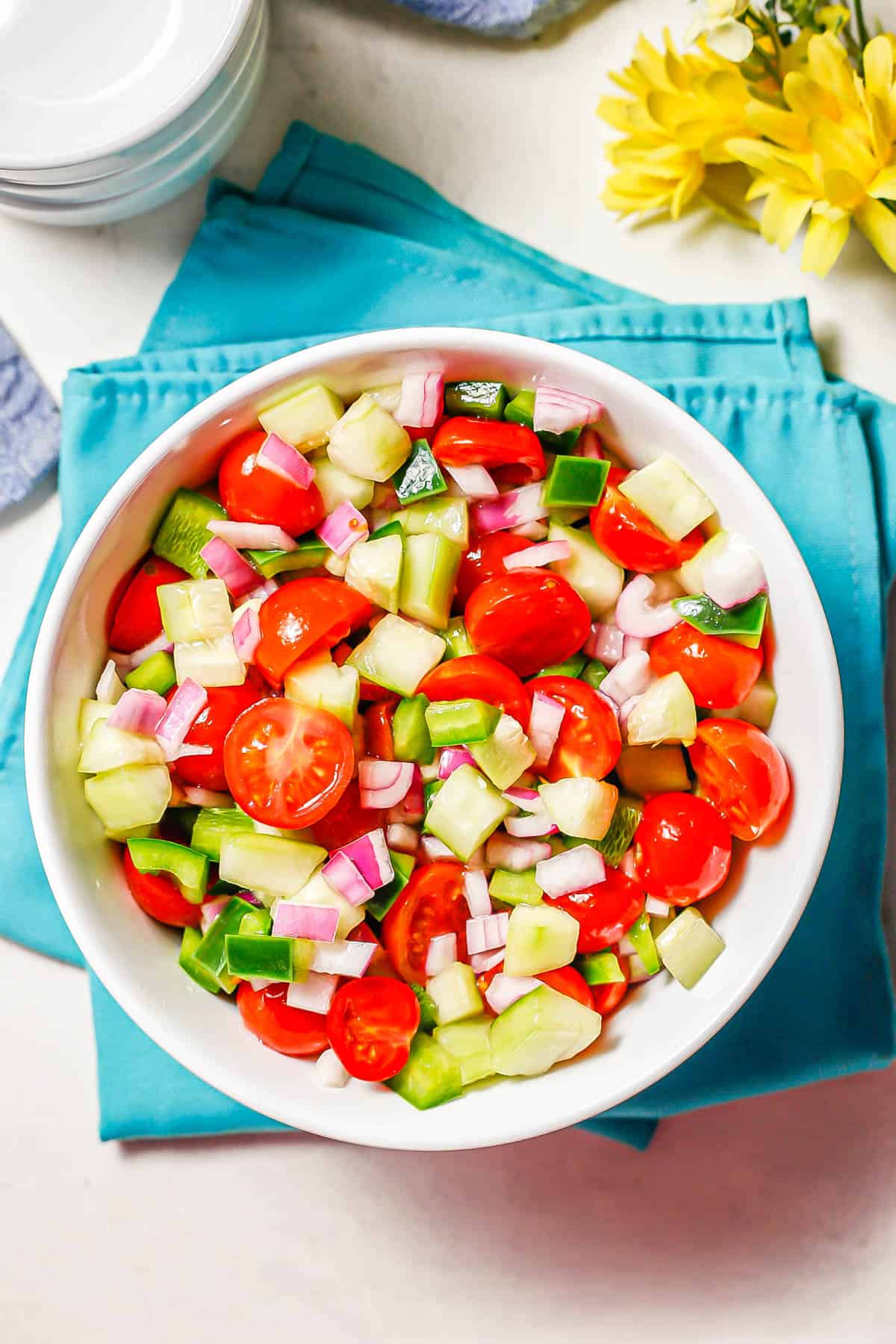 A white serving bowl with a colorful marinated veggie salad set on teal napkins with yellow flowers to the side.