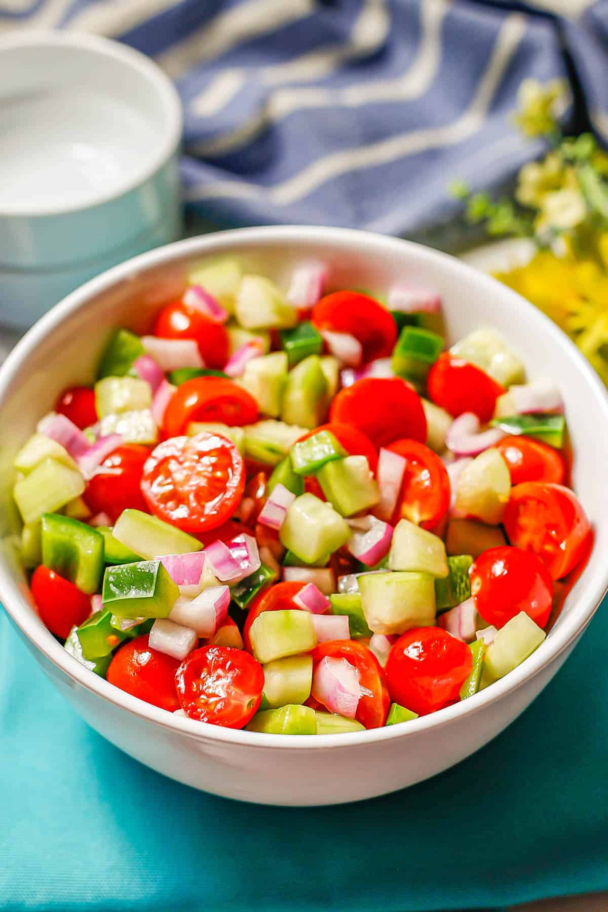 A colorful, fresh vegetable salad in a white bowl set on teal napkins with small white bowls in the background for serving.