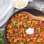 A large skillet with a taco turkey and potato mixture with tomatoes, cilantro and sour cream on top and a bowl of shredded cheese to the side.