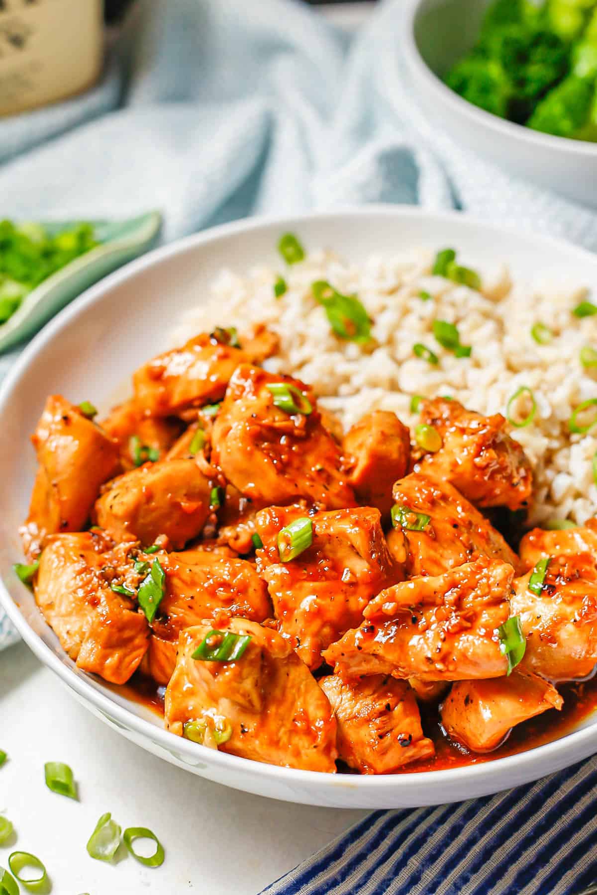 Close up of a saucy bourbon chicken over steamed rice in a white bowl with green onions on top and scattered nearby.