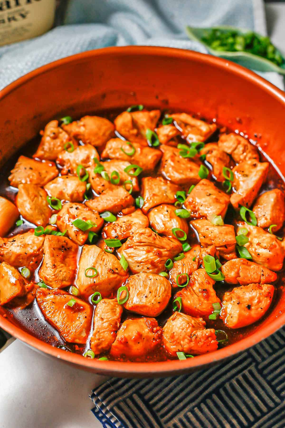 Bourbon chicken in a large copper skillet with sliced green onions on top and a bottle of whiskey in the background.