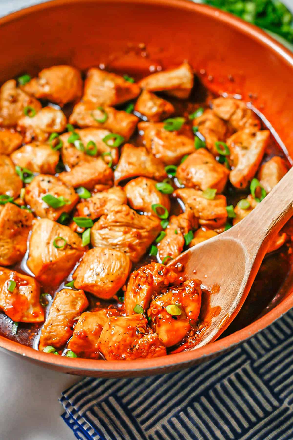 A wooden spoon resting in a large copper skillet with cooked chicken pieces in a dark sauce.