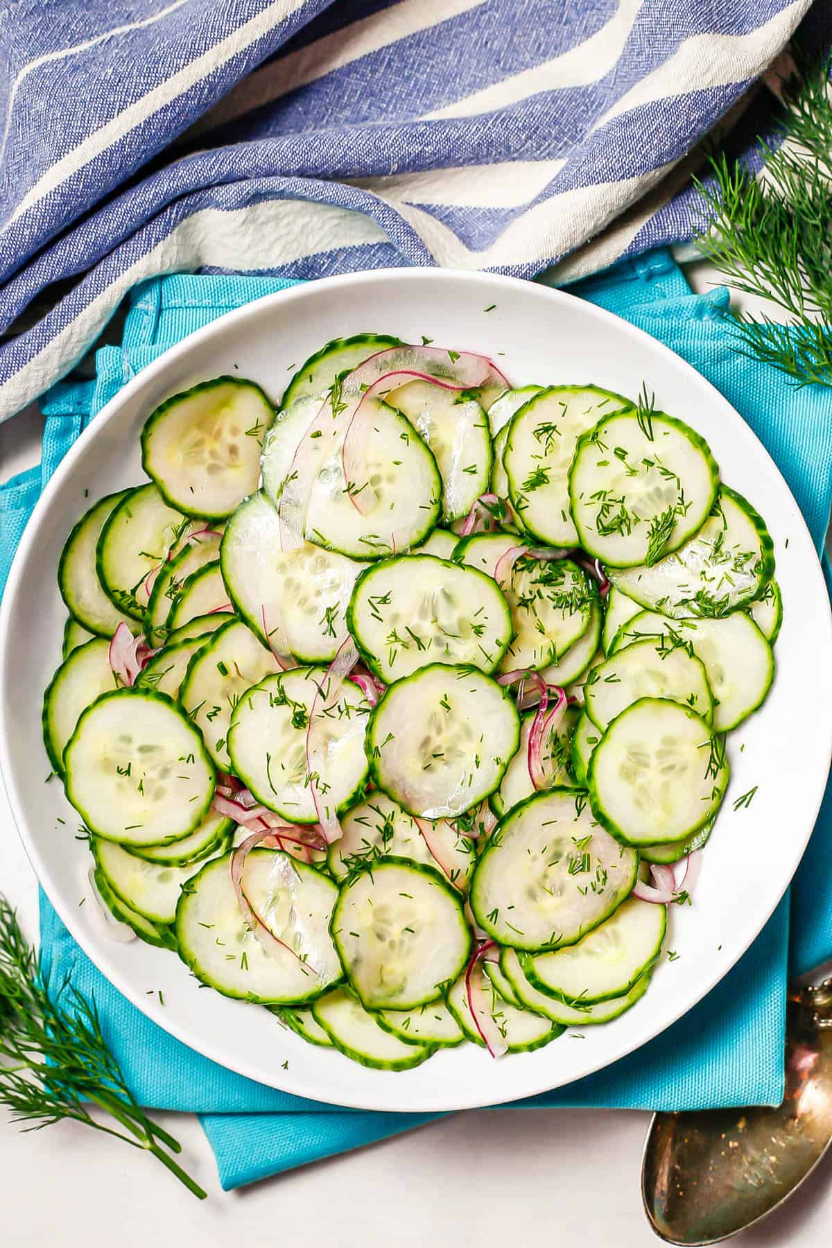 Cucumber salad with fresh dill sprinkled on top served in a low white bowl set on teal napkins.