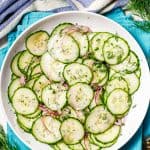 Cucumber salad with fresh dill sprinkled on top served in a low white bowl set on teal napkins with a text overlay on the photo.
