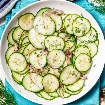 Close up of cucumber salad with fresh dill sprinkled on top served in a low white bowl set on teal napkins.
