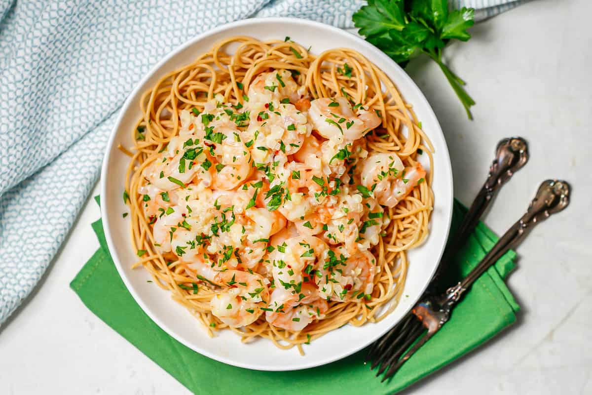 A low white bowl with spaghetti topped with garlic butter shrimp and sprinkled with parsley with two forks to the side.