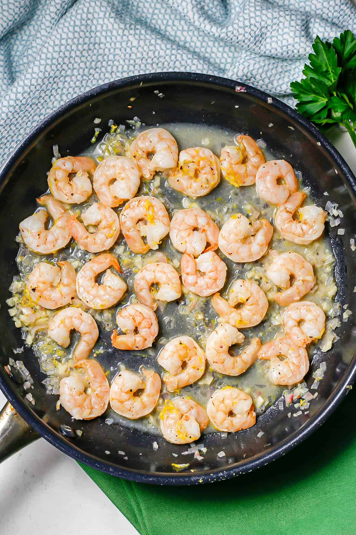 A large skillet with shrimp in a lemony sauce with lemon zest on top and parsley sprigs to the side.