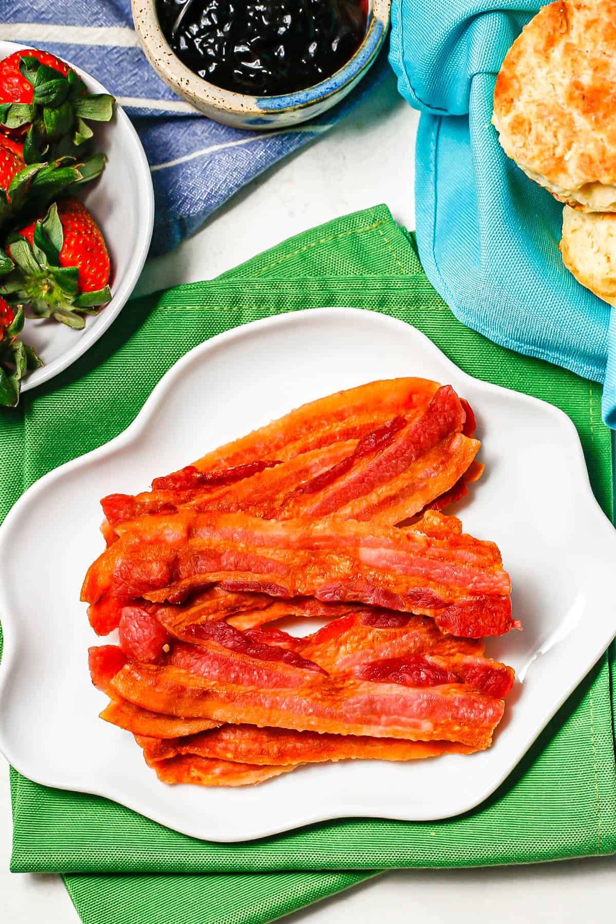 Strips of cooked bacon served on a white plate set on green napkins with fruit and biscuits nearby.