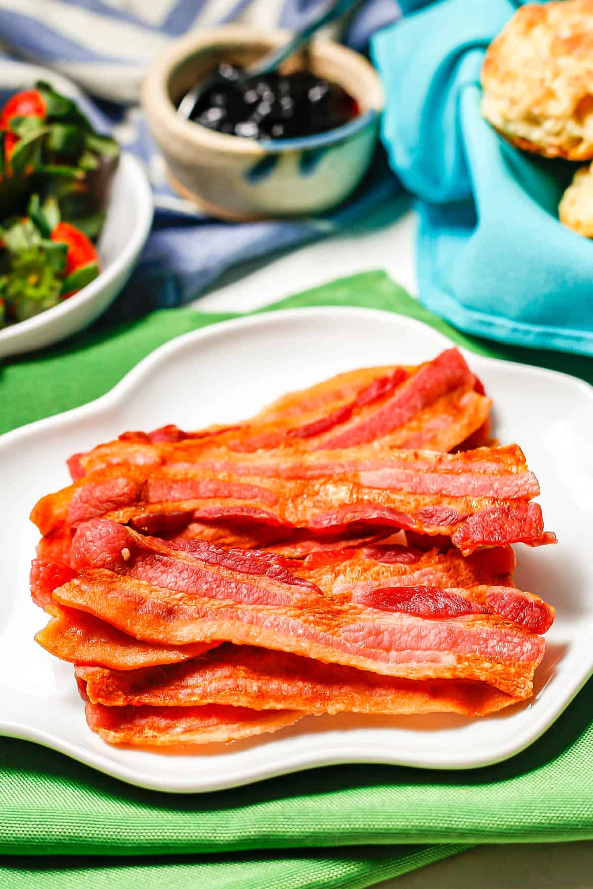 A stack of strips of cooked bacon served on a white plate set on green napkins with fruit and biscuits nearby.