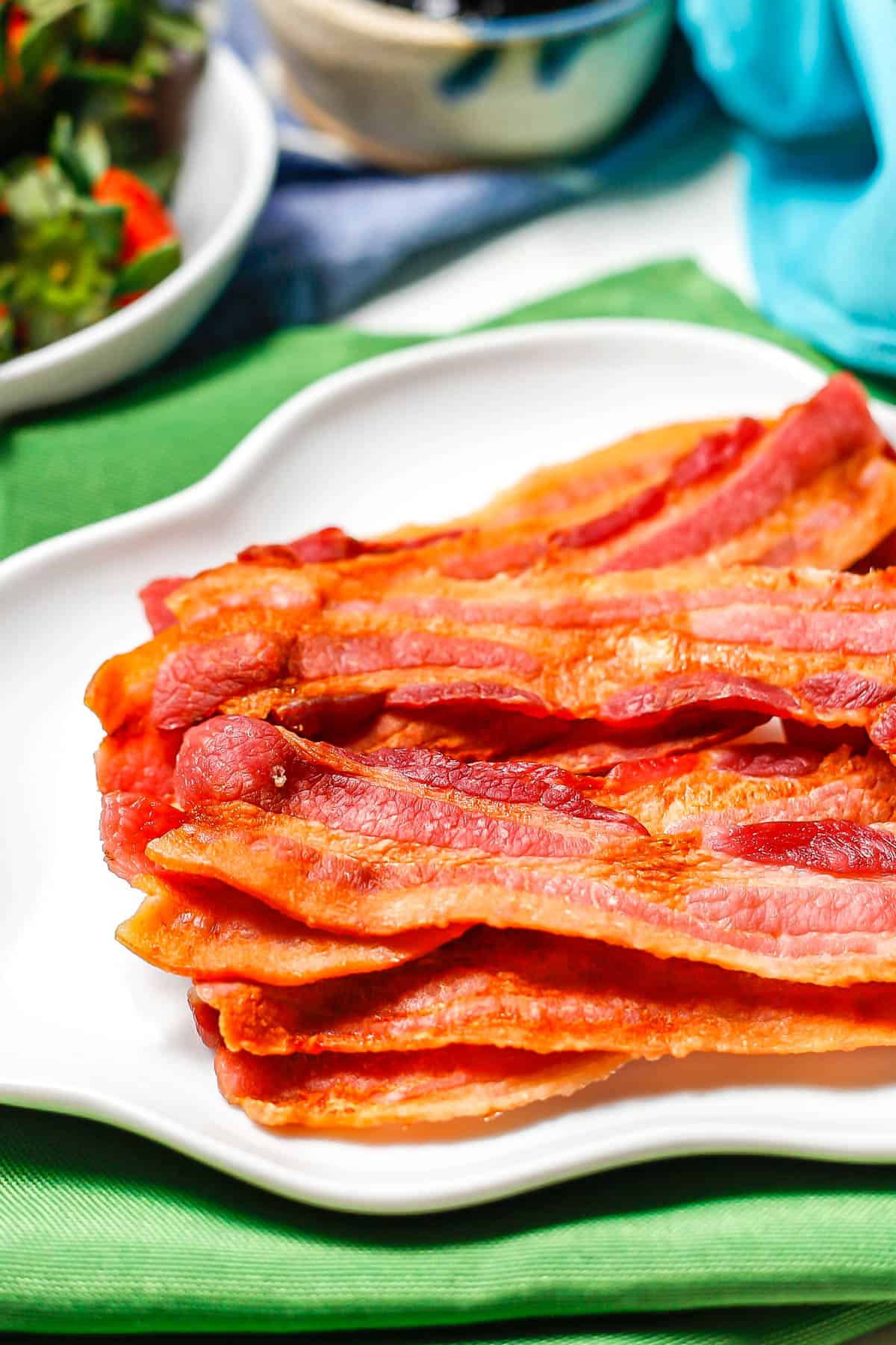 Close up of a stack of cooked bacon strips on a white plate with fluted edges.