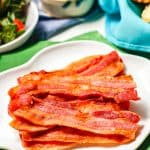 A stack of strips of cooked bacon served on a white plate set on green napkins with fruit and biscuits nearby and a text overlay on the photo.