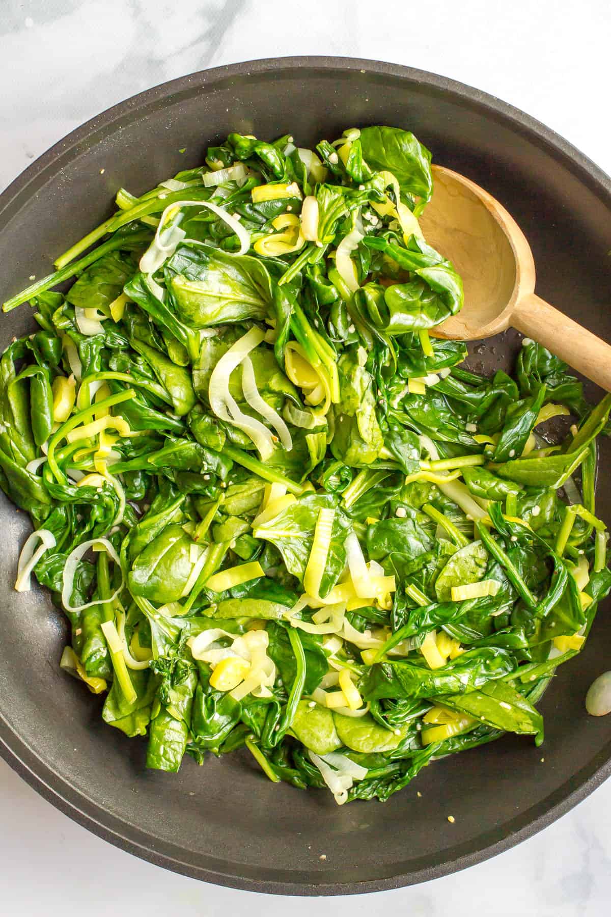 A wooden spoon scooping up some spinach and leeks from a large dark skillet.
