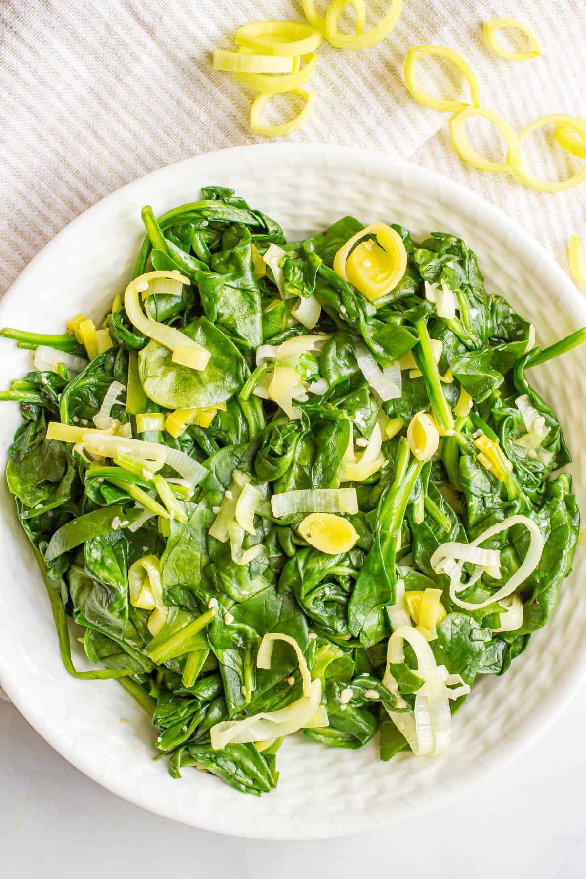 Sautéed spinach and leeks served in a white decorative bowl with sliced leeks in the background.