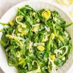 Sautéed spinach and leeks served in a white decorative bowl with sliced leeks in the background and a text overlay on the photo.