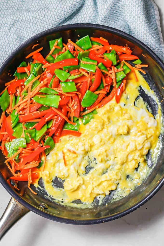 A large skillet with sautéed veggies on one side and eggs being scrambled on the other side.