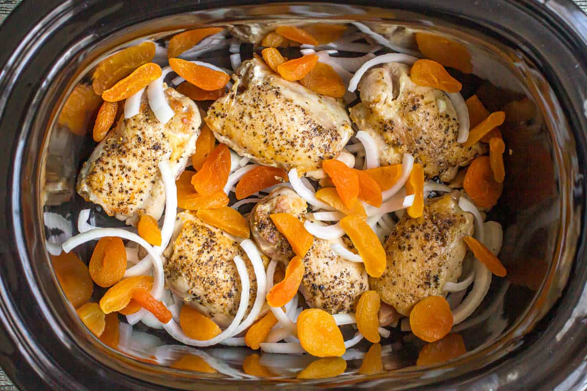 Seared chicken thighs with onions and dried apricots in a slow cooker insert.