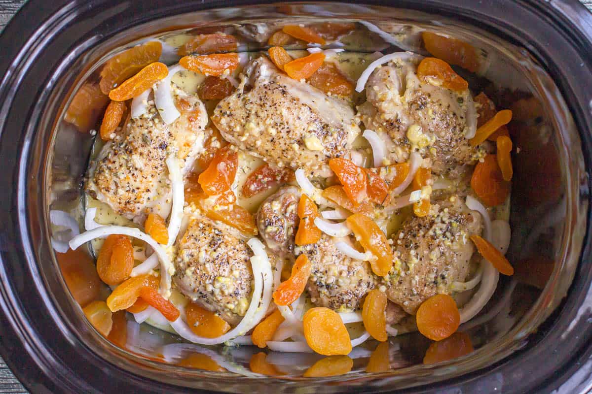 Cooked chicken thighs with onions and dried apricots in a sauce in a slow cooker insert.