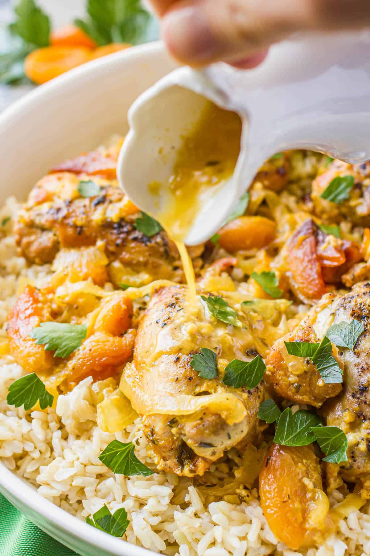 A hand pouring a sauce from a white pitcher over a chicken thigh served on rice in a large white platter.