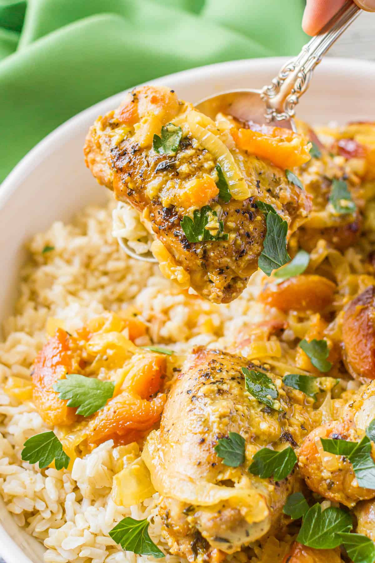 A serving spoon lifting up a chicken thigh with onions and apricots and fresh parsley from a serving dish.