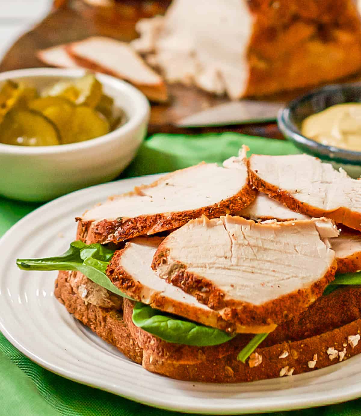 Thick sliced turkey breast layered on a sandwich with spinach underneath and bowls of pickles and mustard in the background.