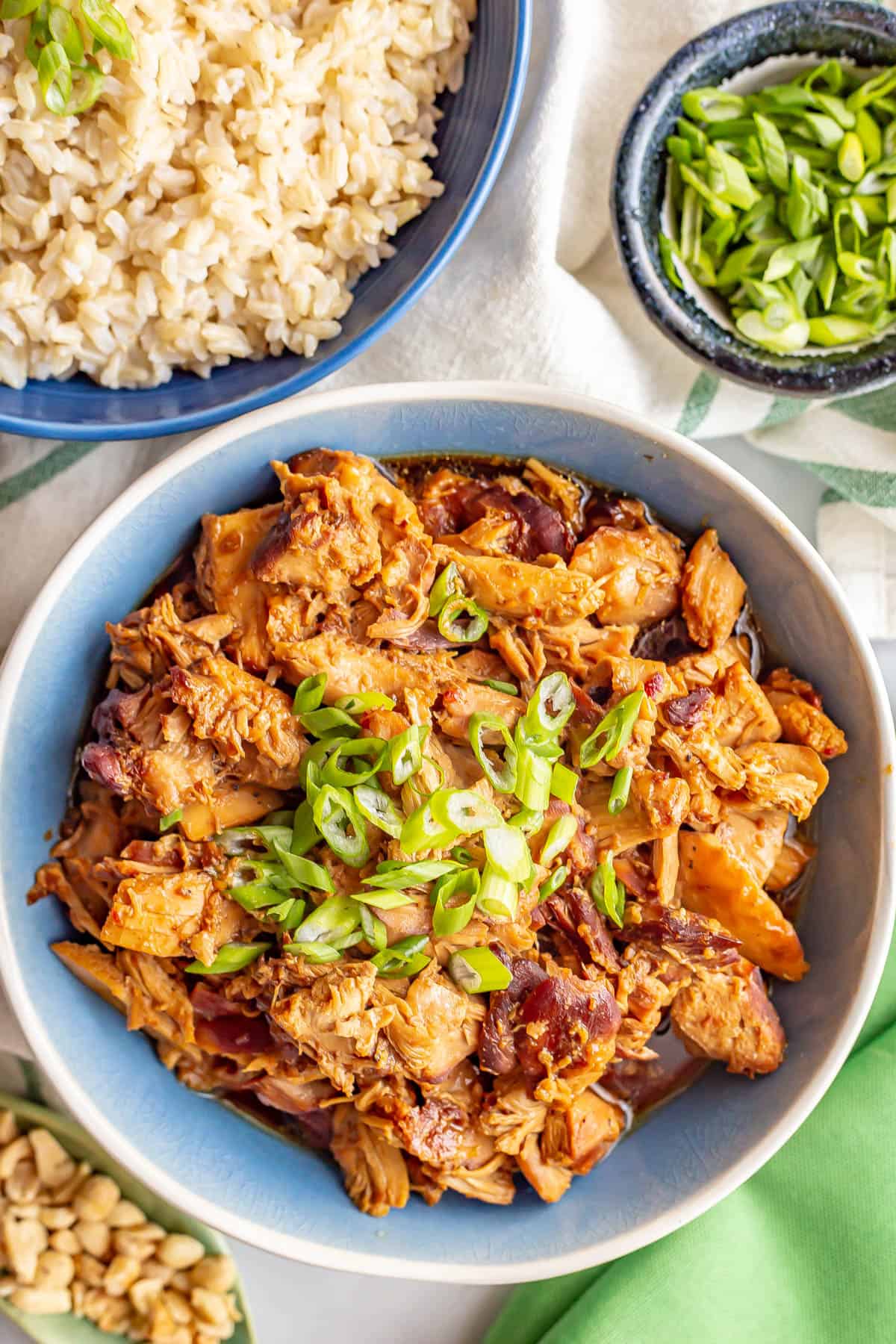 Slow cooker honey garlic chicken served in a bowl with green onions on top with green onions and peanuts in bowls to the side for garnish and a big bowl of rice in the background.