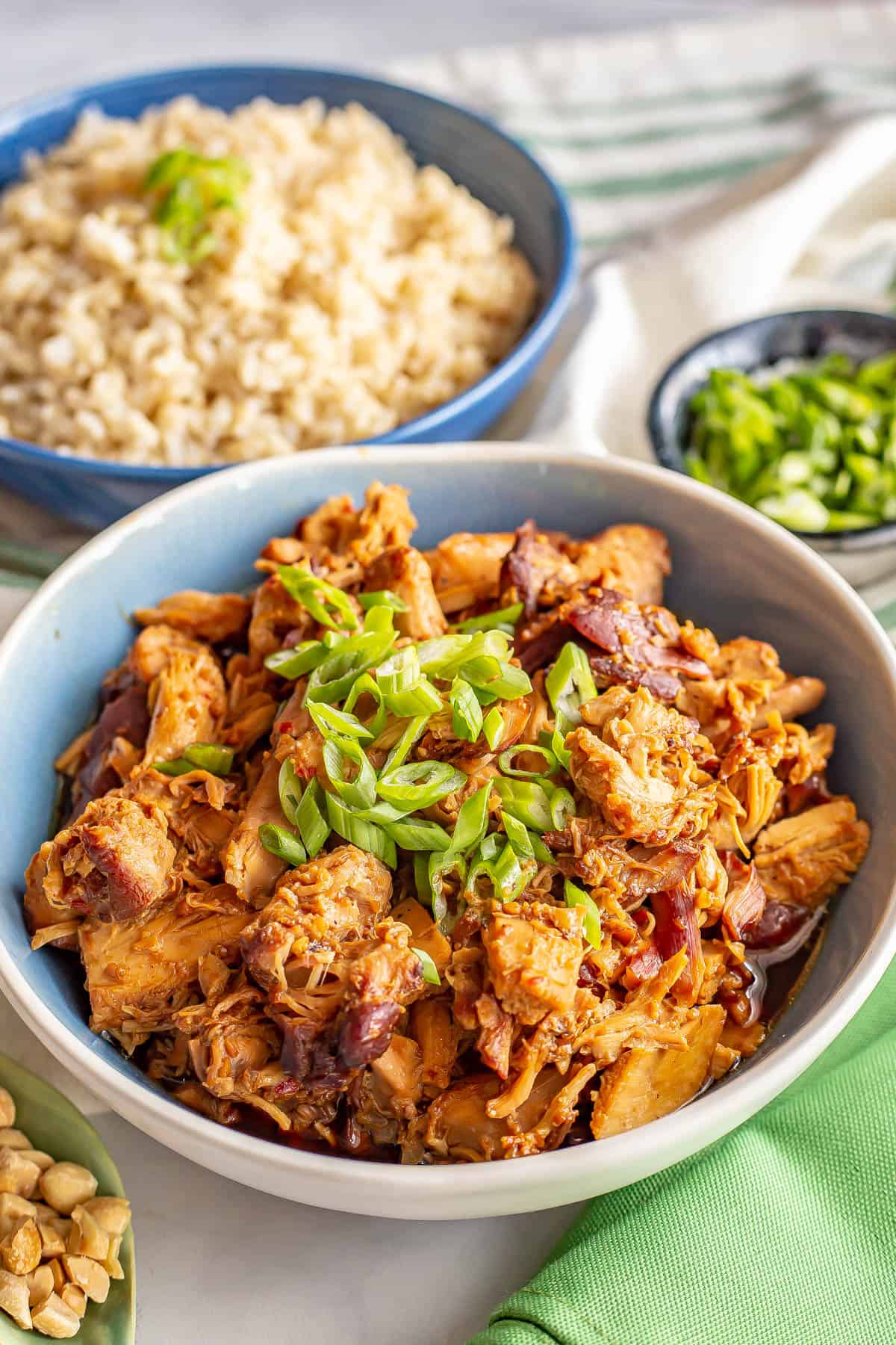 Slow cooker chicken in a honey garlic sauce served in a blue and white bowl with green onions for a garnish and a bowl of steamed rice in the background.