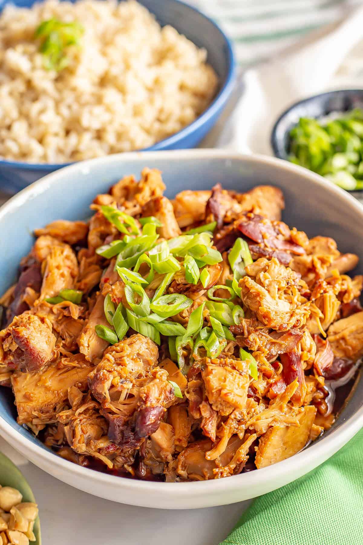 Close up of slow cooker chicken in a honey garlic sauce served in a blue and white bowl with green onions for a garnish and a bowl of steamed rice in the background.