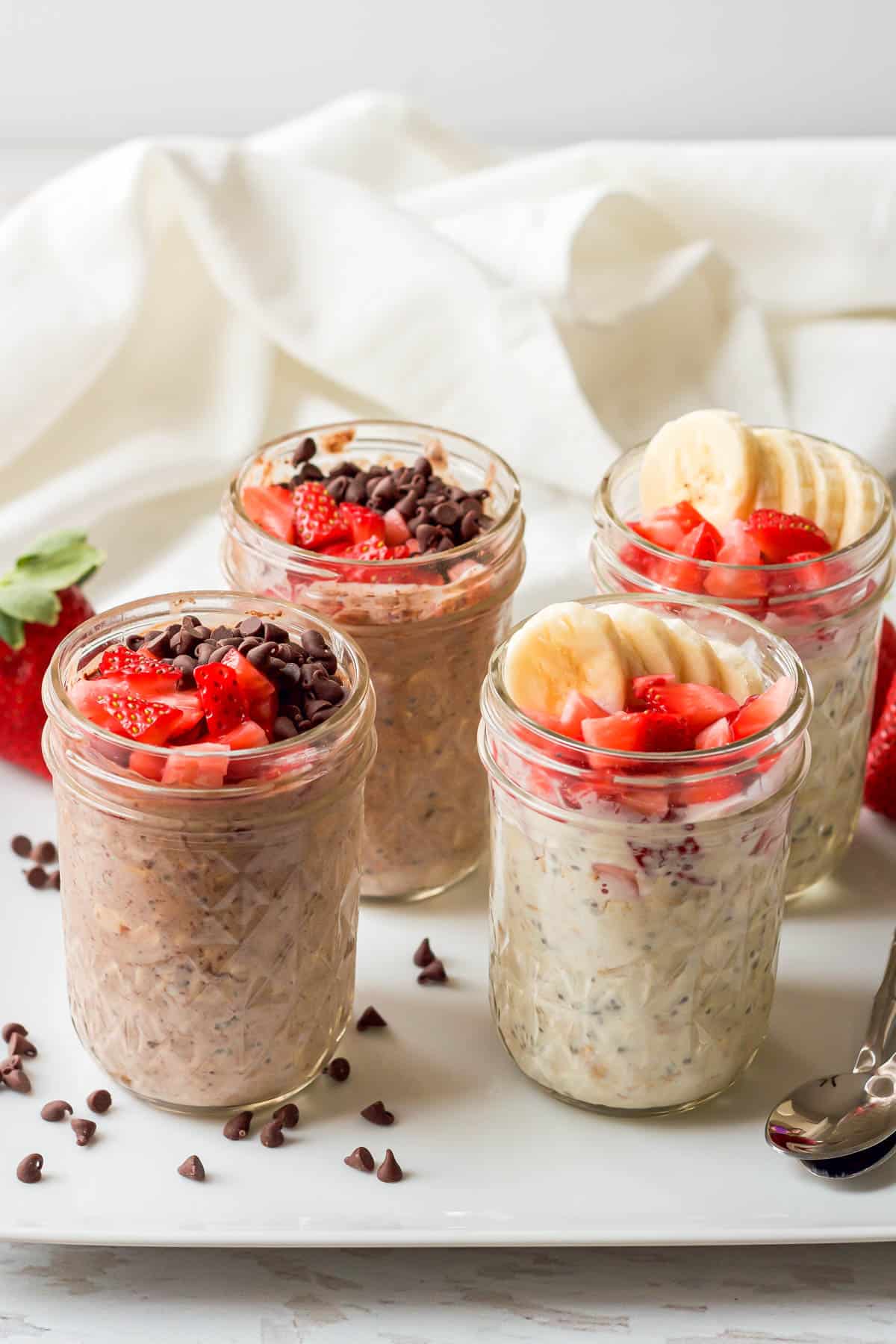 Strawberry overnight oats made 2 different ways and served in small glass jars on a white platter with spoons to the side.