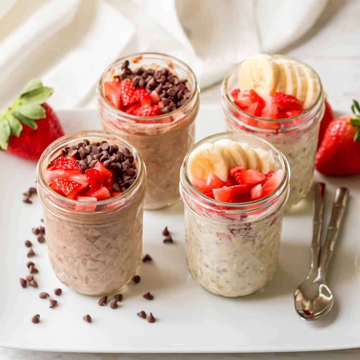 Four small glass jars with strawberry overnight oats made in 2 different ways, with toppings at the top of the jars and on the plate.