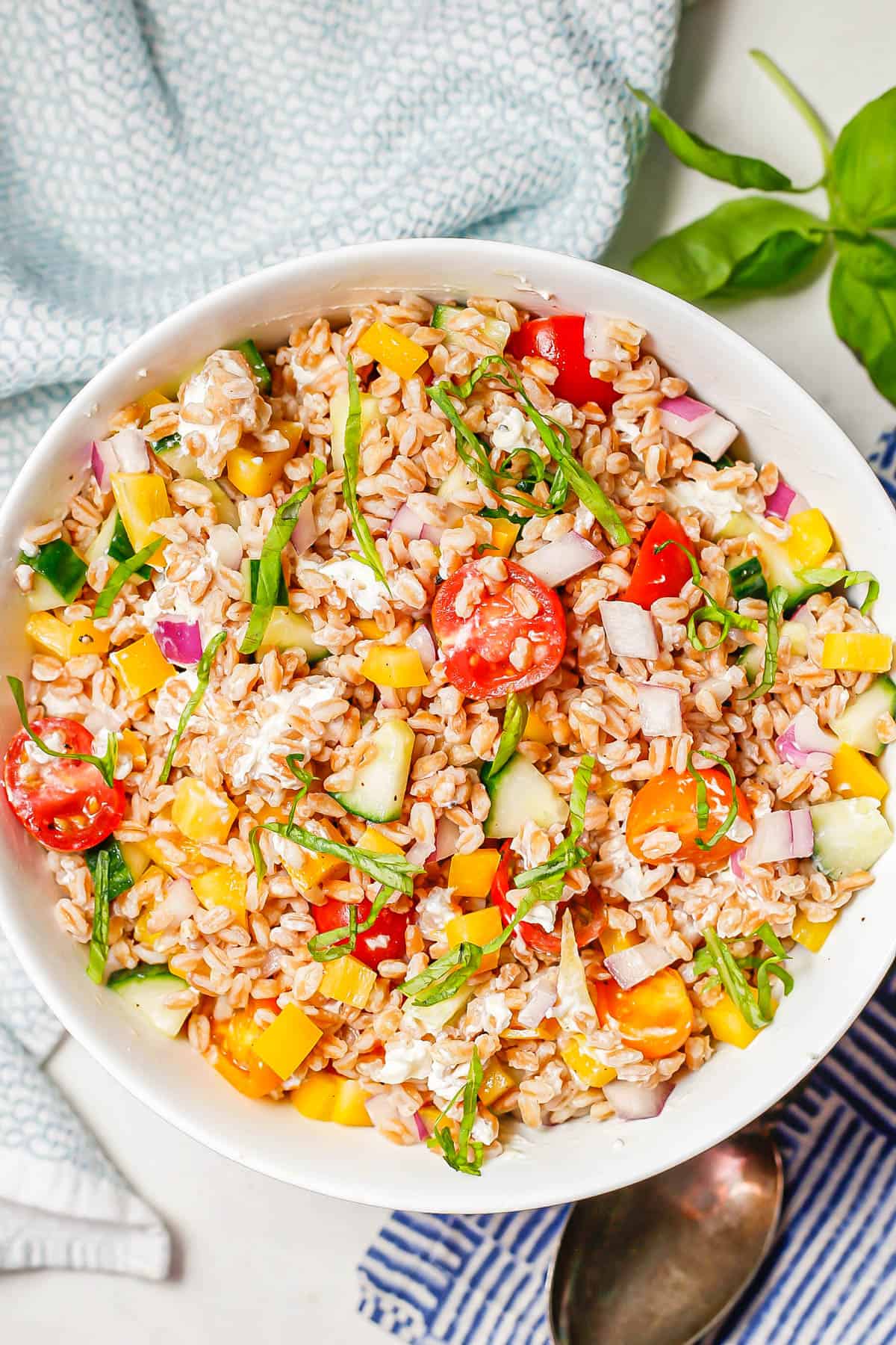 A large white bowl with colorful veggies, farro, goat cheese and basil and a serving spoon to the side.